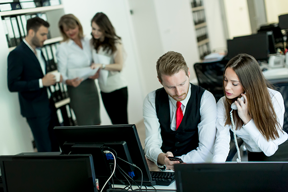 Office professionals working together at computer desks while colleagues collaborate in the background.