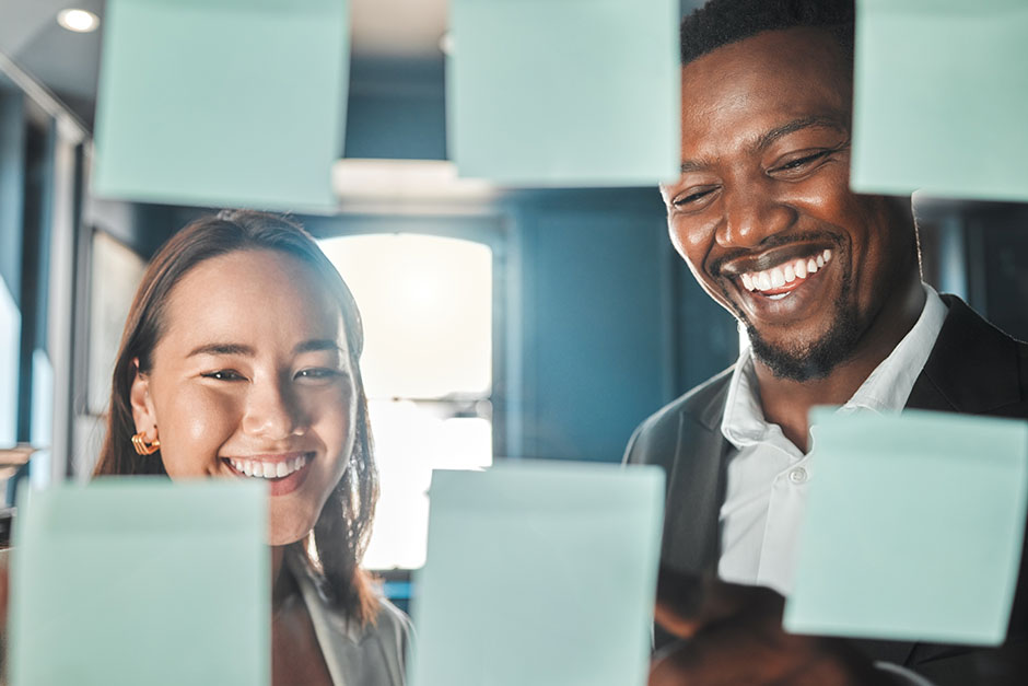 Two smiling professionals brainstorming ideas with sticky notes on a glass wall